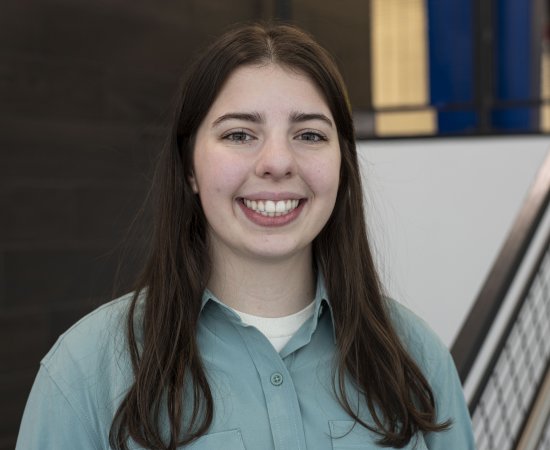 Sydney Kinsella, with long dark hair and wearing a blueish-green button-down top, looks at the camera and smiles.